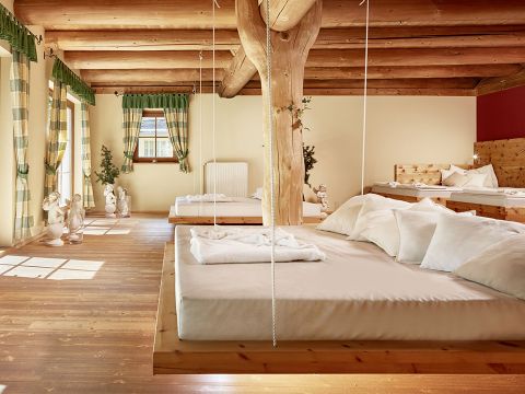 Geräumiges, rustikales Zimmer mit Holzbalkendecke und einem großen hängenden Holzbett mit weißer Bettwäsche. Im Hintergrund stehen mehrere kleine Betten. Sonnenlicht strömt durch Fenster mit grünkarierten Vorhängen und beleuchtet den Holzboden und das Dekor.