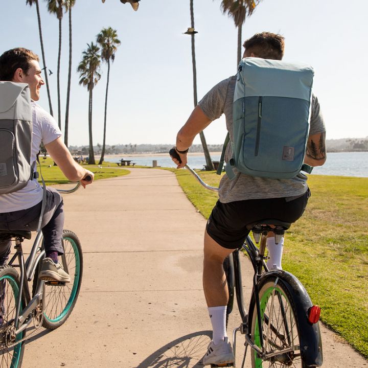 Zwei Personen fahren mit dem Fahrrad auf einem gepflasterten Weg entlang eines Parks am Wasser. Beide tragen Rucksäcke und legere Kleidung. Auf beiden Seiten des Weges sind Palmen und Gras, im Hintergrund ist das Wasser unter einem blauen Himmel zu sehen.