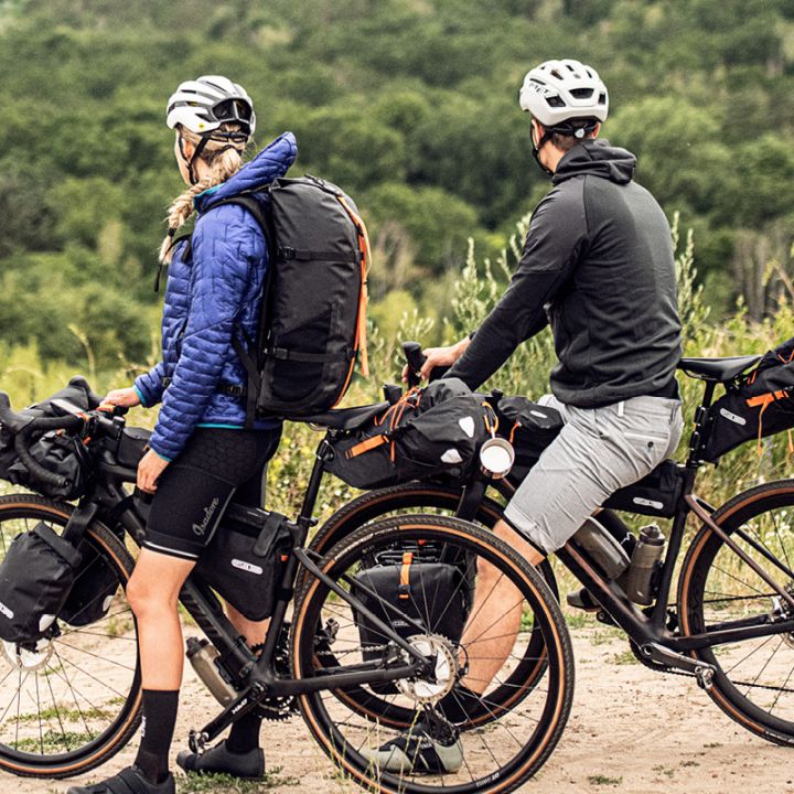 Zwei Radfahrer mit Helmen und Outdoor-Ausrüstung stehen mit ihren beladenen Tourenrädern auf einem Feldweg. Ihr Blick geht auf einen üppigen, grünen Wald. Beide Fahrräder sind mit mehreren Taschen für die Ausrüstung ausgestattet.