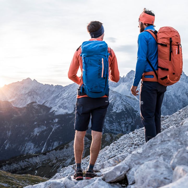 Zwei Wanderer mit Rucksäcken stehen an einem felsigen Berghang und betrachten einen fernen Sonnenuntergang über schroffen Gipfeln. Der linke Wanderer trägt eine blaue Jacke, während der rechte Wanderer einen orangefarbenen Hut und einen Rucksack trägt. Die Landschaft darunter ist im Schatten.