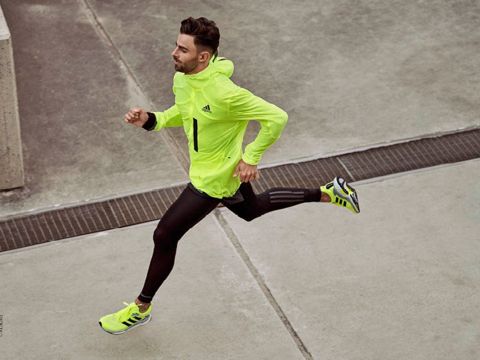 Eine Person in leuchtend gelbgrüner Sportbekleidung und schwarzen Leggings läuft im Freien. Die Szene zeigt Bewegung, mit Betonumgebung und einem Metallgitter im Hintergrund.