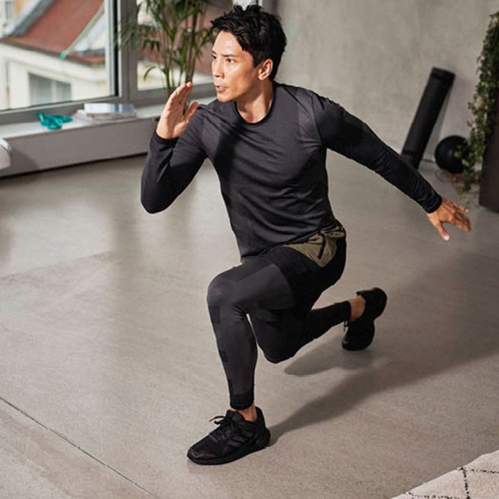 Eine Person in Sportkleidung führt in einem Raum eine Ausfallschrittübung durch. Ein Knie ist gebeugt, das andere Bein ist nach hinten ausgestreckt, die Arme sind so positioniert, als ob sie in Bewegung wären. Der Raum hat große Fenster, einen hellen Boden und im Hintergrund Trainingsgeräte.