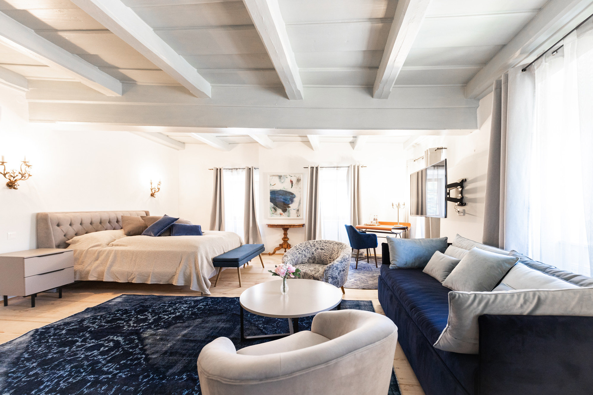 Ein geräumiges, gut beleuchtetes Schlafzimmer mit weißen Wänden und freiliegenden Holzbalken. Es verfügt über ein Bett, ein blaues Sofa, einen Sessel und einen runden Couchtisch. Ein Teppich verleiht Struktur und große Fenster lassen natürliches Licht in den Raum. Im Hintergrund sind ein Gemälde und ein Schreibtisch zu sehen.