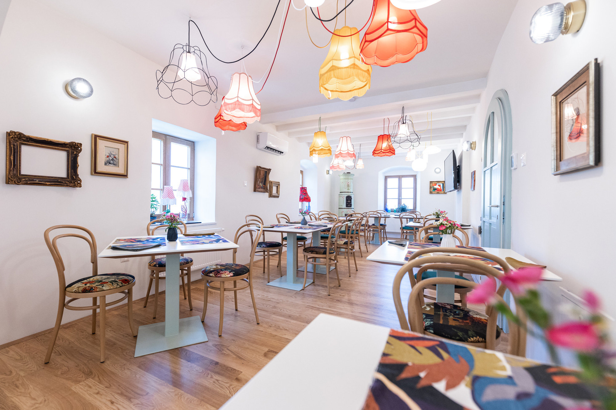 Ein gemütliches Café mit Holzboden, dekoriert mit bunten Hängelampen und hübsch gerahmten Bildern. Die Tische und Stühle sind mit Blumenmustern verziert und Blumenvasen schmücken die Tische. Große Fenster lassen den Raum mit natürlichem Licht durchfluten.