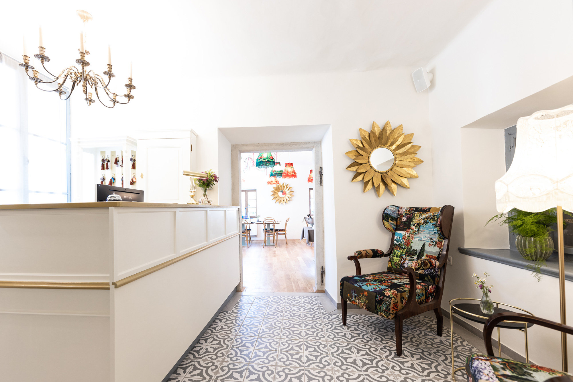 Das helle und gemütliche Interieur verfügt über eine Rezeption mit Blumendekor, einen gemusterten Sessel, einen Sonnenspiegel und bunte Fliesen. Eine Tür führt zu einem Essbereich und von der Decke hängt ein Kronleuchter.