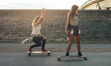 Zwei Personen fahren auf Longboards auf einer asphaltierten Straße neben einer Steinmauer. Einer balanciert mit erhobenen Armen, während der andere in legerer Kleidung mit um die Hüften gebundenen Hemden vorwärts gleitet. Im Hintergrund geht die Sonne unter.