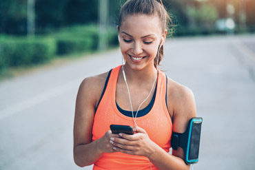 Eine Frau in einem leuchtend orangefarbenen Tanktop joggt im Freien. Sie trägt Kopfhörer und lächelt, während sie auf ihr Smartphone schaut, das sie in einer Hand hält. An ihrem anderen Arm ist eine Handyhalterung befestigt. Hinter ihr sind ein verschwommener Pfad und Grün zu sehen.