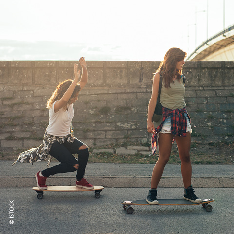 Zwei Personen fahren an einem sonnigen Tag Longboard. Einer hockt und hebt die Arme, während der andere auf seinem Board steht und nach vorne schaut. Beide tragen legere Kleidung und haben ihre Hemden um die Hüften gebunden. Im Hintergrund ist eine Steinmauer zu sehen.