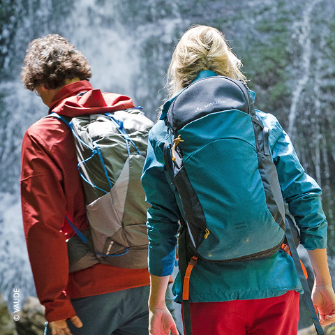 Zwei Personen mit Rucksäcken wandern in der Nähe eines Wasserfalls. Sie tragen Outdoor-Kleidung, einer trägt eine rote Jacke und der andere eine blaue Jacke. Die Szene vermittelt ein Gefühl von Abenteuer und Naturerkundung.