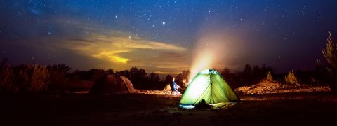 Eine Nachtszene mit zwei Zelten in einer Wüstenlandschaft unter einem Sternenhimmel. Ein Zelt ist von innen beleuchtet und verbreitet ein warmes Licht. In der Nähe brennt ein Lagerfeuer und schafft eine gemütliche Atmosphäre mit Baumsilhouetten im Hintergrund.