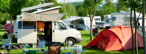 Ein Campingplatz mit einem Wohnmobil und einem orangefarbenen Zelt. Das Seitenvordach des Wohnmobils ist ausgefahren. Im Vordergrund stehen ein Tisch mit Stühlen und Campingausrüstung. Im Hintergrund sind mehrere andere Wohnmobile zwischen Bäumen geparkt.