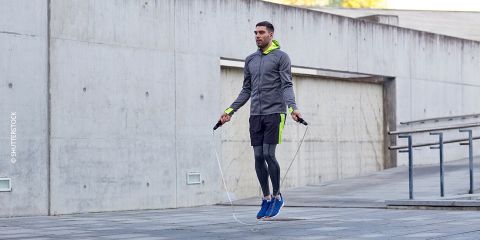 Ein Mann springt Seil auf einer Betonfläche. Er trägt einen grauen Kapuzenpullover mit Neonakzenten, schwarze Shorts, Leggings und blaue Turnschuhe. Im Hintergrund sind eine Betonwand und ein Geländer zu sehen.