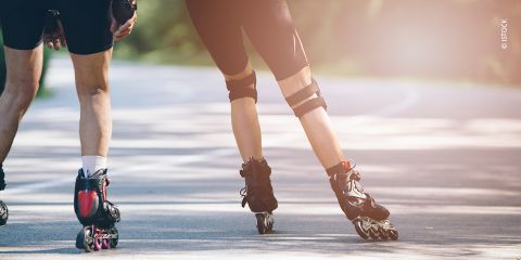 Zwei Personen fahren auf Rollschuhen auf einem glatten Weg, umgeben von Grün. Der Fokus liegt auf ihren Unterkörpern, die Sportbekleidung und Rollschuhe zeigen. Sonnenlicht wirft weiche Schatten auf den Bürgersteig.