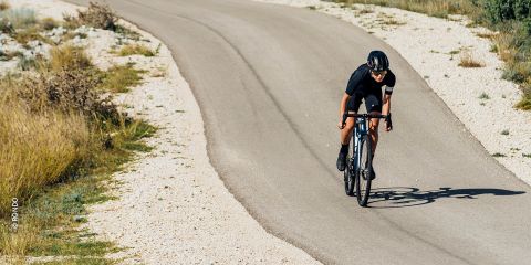 Ein Radfahrer in schwarzem Outfit und Helm fährt mit seinem Rennrad auf einem kurvenreichen, gepflasterten Weg durch eine trockene, felsige Landschaft. Die Sonne wirft Schatten auf die Straße, als sich der Radfahrer in eine Kurve legt.