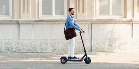 Eine Person mit Bart und Brille fährt mit einem Elektroroller eine Straße entlang. Sie trägt ein blaues Hemd, weiße Hosen und eine braune Tasche. Sie kommt an einem Gebäude mit großen Fenstern und einer beigen Fassade vorbei.