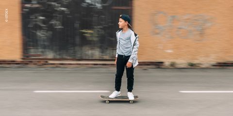 Ein kleiner Junge mit einer Kappe fährt mit Skateboard eine Straße entlang. Er trägt einen Kapuzenpullover, ein T-Shirt und Jeans. Im Hintergrund ist eine verschwommene Hauswand mit Graffiti zu sehen.