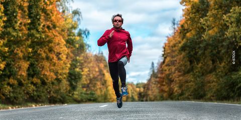 Eine Person in einer roten Jacke und schwarzen Leggings läuft auf einer leeren Straße, umgeben von Bäumen mit Herbstlaub. Sie trägt eine Sonnenbrille und der Himmel ist teilweise bewölkt.