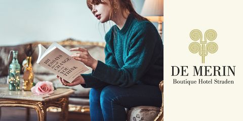 Eine Frau mit roten Haaren, einem grünen Pullover und Jeans, sitzt auf einem Sofa und liest ein Buch mit dem Titel „101 Essays, die Ihre Denkweise verändern werden“. Auf einem Couchtisch neben ihr stehen eine rosa Rose und eine Glasflasche. Rechts das Logo des De Merin Boutique Hotel, Straden.