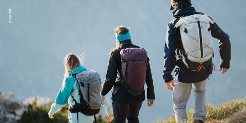 Drei Wanderer, mit Jacken und Rucksäcken bekleidet, gehen auf einem grasbewachsenen Bergpfad mit dunstigen blauen Bergen im Hintergrund. Die Szene vermittelt ein Gefühl von Abenteuer und Erkundung in der Natur.