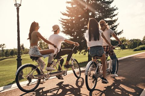 Vier junge Erwachsene fahren auf einem sonnigen Parkweg Tandemfahrräder. Je zwei Paare teilen sich ein Fahrrad und genießen die Natur. Die Gruppe ist leger gekleidet und das Sonnenlicht fällt durch die Bäume und verleiht der Szene warme Farbtöne.