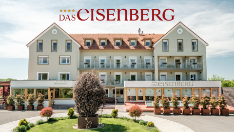 Vorderansicht eines mehrstöckigen Gebäudes mit Balkonen, umgeben von Grünanlagen und Topfpflanzen. Auf der oberen Fassade prangt ein großes Logo mit „Das Eisenberg“. Die Umgebung ist sonnig und gepflegt und vermittelt eine einladende Atmosphäre.