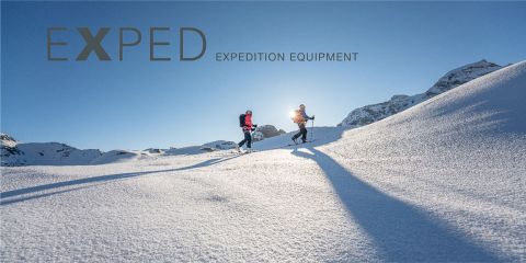 Zwei Personen in Winterkleidung laufen durch eine schneebedeckte Landschaft, während das helle Sonnenlicht lange Schatten wirft. Im Hintergrund sind Berge zu sehen. Oben links steht der Text „EXPED EXPEDITION EQUIPMENT“.