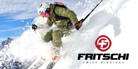 Ein Skifahrer in grünem Outfit und weißem Helm fährt durch tiefen Schnee und wirbelt dabei Pulverschnee auf. Im Hintergrund ist der Himmel klar und blau. Das Bild zeigt das Logo und den Text: „Fritschi Swiss Bindings“.