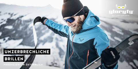 Eine Person in blauer Jacke und Skibrille hält Skier vor einer schneebedeckten Bergkulisse. Der Text lautet „Unzerbrechliche Brillen“ und „Produziert in Österreich“. In der oberen rechten Ecke befindet sich ein Logo.