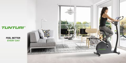 Eine Person trainiert auf einem Heimtrainer in einem modernen Wohnzimmer mit großen Fenstern und Blick auf eine grüne Landschaft. Der Raum verfügt über ein graues Sofa, gemusterte Kissen, einen hellen Teppich und eine elegante Einrichtung. Der Text lautet: „Tunturi, fühle dich jeden Tag besser.“.