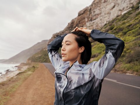 Eine Person in einer grauen Jacke steht an einer Küstenstraße und richtet ihre Haare. Sie befindet sich in der Nähe einer Felsklippe. Links ist das Meer zu sehen. Der Himmel ist bewölkt, was eine stimmungsvolle Atmosphäre schafft.