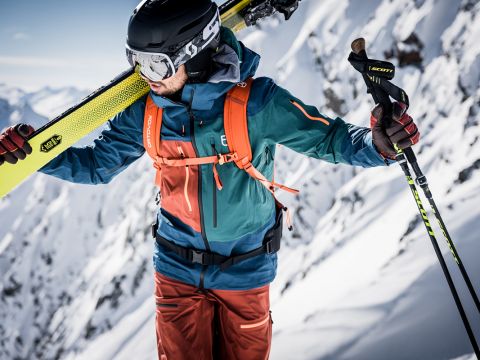 Ein Skifahrer in einer bunten Jacke und Hose trägt Skier auf der Schulter. Er hält einen Skistock und trägt einen Helm und eine Schutzbrille. Der Hintergrund zeigt eine schneebedeckte Bergkette unter einem klaren blauen Himmel.
