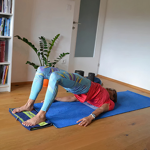 Eine Person in einem roten Hemd und gemusterten Leggings macht eine Brückenpose auf einer blauen Matte in einem hellen Raum. Im Hintergrund sind eine Pflanze und Bücherregale zu sehen.