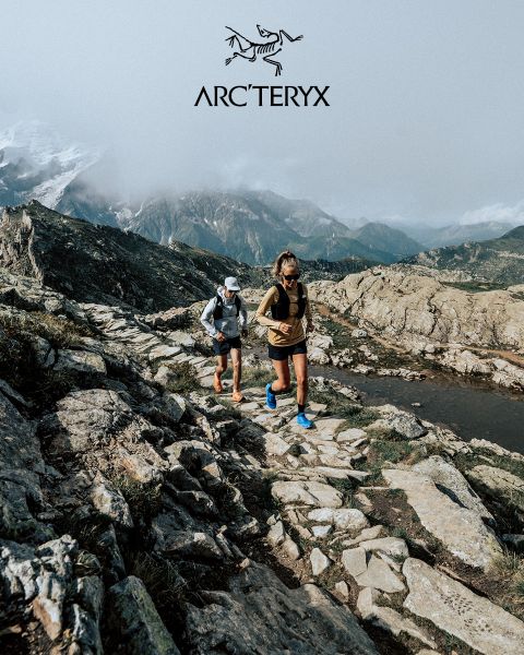 Zwei Personen wandern auf einem felsigen Bergpfad. Der Himmel ist bedeckt, im Hintergrund ist eine neblige Bergkette zu sehen. Beide Wanderer tragen Outdoor-Ausrüstung. Rechts befindet sich ein kleiner Teich. Oben ist das Arc'teryx-Logo zu sehen.