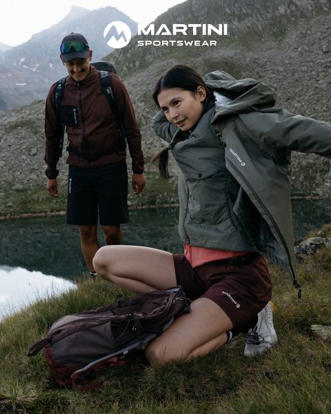 Zwei Personen in Outdoor-Ausrüstung mit Rucksäcken stehen an einem Bergsee. Eine kniet und richtet ihre Jacke, die andere steht lächelnd da. Im Hintergrund felsiges Gelände und bewölkter Himmel. Oben im Bild ist „MARTINI SPORTSWEAR“ zu sehen.