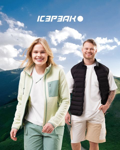 Eine lächelnde Frau und ein lächelnder Mann posieren im Freien vor einer Berglandschaft im Hintergrund. Die Frau trägt eine hellgrüne Jacke und Hose, der Mann ein weißes Hemd, eine schwarze Weste und beige Shorts. Beide stehen unter einem blauen Himmel mit dem „Icepeak“-Logo darüber.