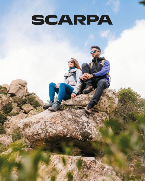 Eine Frau und ein Mann sitzen im Freien auf großen Felsen. Sie tragen Freizeitkleidung und Sonnenbrillen. Sie sind von Grün und Felsen umgeben, der Himmel ist leicht bewölkt. Oben im Bild ist das Wort „SCARPA“ zu sehen.