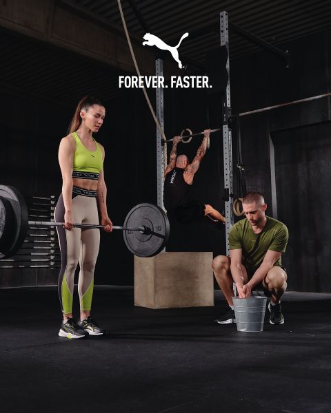 Eine Frau in Sportkleidung stemmt eine Hantel, ein Mann hockt mit einem Eimer neben ihr, eine weitere Person hängt an Ringen in einem Fitnessstudio. Oben steht der Text „FOREVER. FASTER.“ mit einem Puma-Logo.