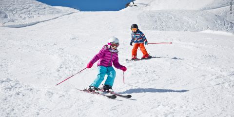 Zwei Kinder fahren auf einem verschneiten Hang Ski. Das Kind vorne trägt eine lila Jacke und hellblaue Hosen und hält rote Stöcke. Das Kind dahinter trägt ein orangefarbenes Outfit mit einer gestreiften Jacke. Beide tragen Helme und Schutzbrillen. Heller, klarer Himmel darüber.