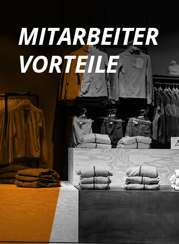 Innenansicht eines Bekleidungsgeschäfts mit ordentlich gefalteter Kleidung auf Regalen und Ständern. Auf einem Schild steht „MITARBEITER VORTEILE“, was auf Mitarbeitervorteile hinweist. Die Kulisse umfasst verschiedene Jacken und Hemden, die vor einem teilweise orangefarbenen Hintergrund ausgestellt sind.