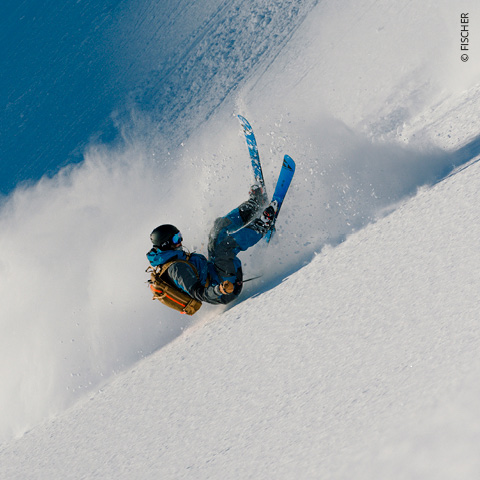Ein Skifahrer in blauer Jacke und Helm fährt einen verschneiten Hang hinunter und wirbelt dabei eine Schneewolke auf. Der Skifahrer neigt sich zur Seite, im Hintergrund ist der blaue Himmel zu sehen.