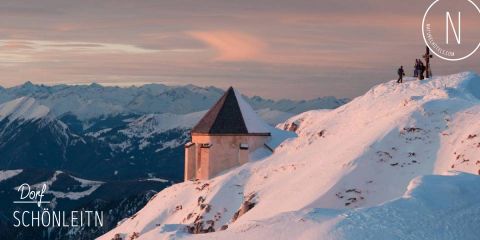 Eine schneebedeckte Berglandschaft bei Sonnenuntergang mit einem kleinen Gebäude mit Spitzdach auf einem Bergrücken. Menschen stehen in der Nähe und bewundern die Aussicht. In der Ferne erstrecken sich schneebedeckte Bergketten unter einem pastellfarbenen Himmel.