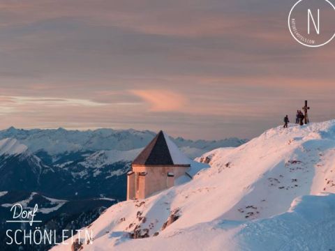Schneebedeckte Berglandschaft bei Sonnenaufgang, mit einem kleinen Gebäude und einem Kreuz auf einem Gipfel. Mehrere Menschen bewundern die Aussicht und in der Ferne erstrecken sich schneebedeckte Berge unter einem farbenfrohen Himmel. Text: „Dorf Schönleiten N/We Live Style.com“ in den Ecken.