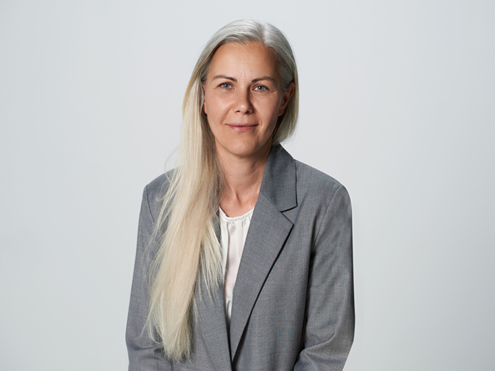 Eine Person mit langem, glattem, hellem Haar trägt einen grauen Blazer und eine weiße Bluse. Sie sitzt da und blickt mit neutralem Gesichtsausdruck vor einem schlichten hellen Hintergrund in die Kamera.