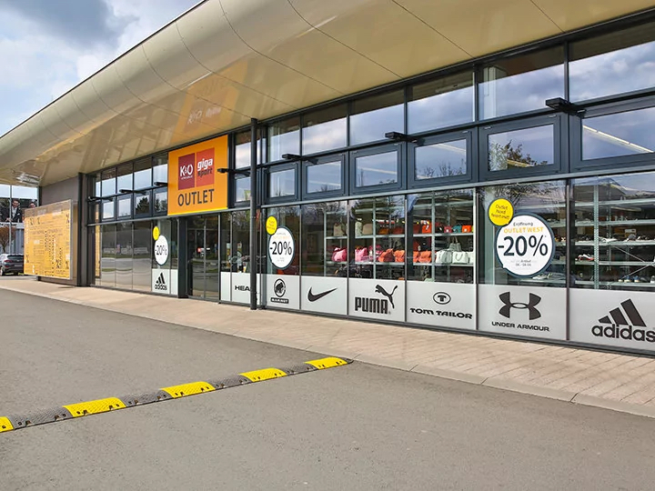 Außenansicht eines Sport-Outlet-Stores mit großen Fenstern, auf denen verschiedene Markenlogos wie Puma und Adidas zu sehen sind. Schilder an den Fenstern werben mit 20 % Rabatt auf Möbel, Vorhänge und Heimtextilien. Das Gebäude besticht durch sein modernes Design.
