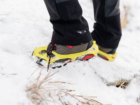 Nahaufnahme einer Person, die Steigeisen über Wanderschuhen trägt und auf Schnee läuft. Die Steigeisen sind gelb mit roten Akzenten und bieten Halt auf der eisigen Oberfläche. Die Person trägt schwarze Hosen und man sieht etwas Gras, das durch den Schnee ragt.