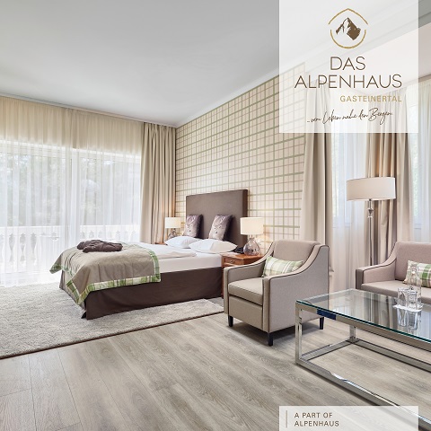 Ein modernes Hotelzimmer mit einem großen Doppelbett, zwei Sesseln, einem Couchtisch aus Glas und einer Stehlampe. Das Zimmer verfügt über eine karierte Akzentwand und ein großes Fenster mit Vorhängen. Das Logo „Das Alpenhaus“ ist auf dem Bild sichtbar.