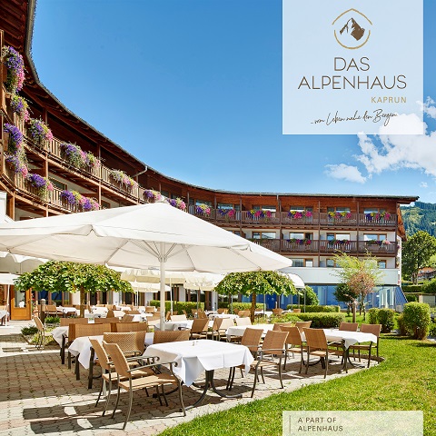 Essbereich im Freien mit Tischen und Stühlen unter Sonnenschirmen, umgeben von einem geschwungenen Gebäude mit blumengeschmückten Balkonen. Der klare blaue Himmel verstärkt die helle, heitere Atmosphäre. Auf dem Schild steht „Das Alpenhaus Kaprun“.