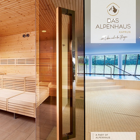 Innenansicht einer Sauna mit Holzbänken und -wänden und einer großen Glastür. Von außen bietet sich durch die großen Fenster ein Blick auf einen Garten und die Berge. Auf dem Glas ist der Text „Das Alpenhaus Kaprun“ zu sehen.