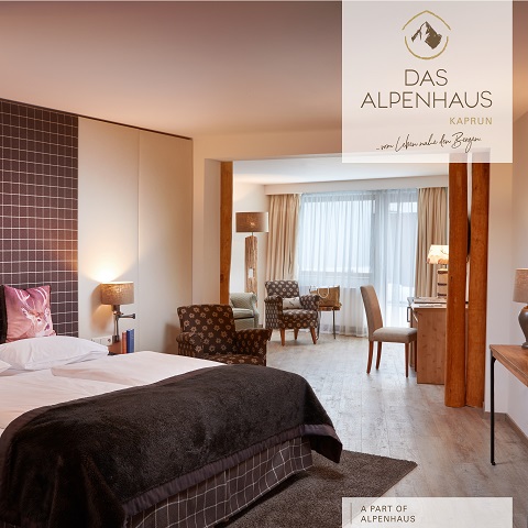 Ein gemütliches Hotelzimmer mit einem großen Bett, braunen Decken und dekorativen Kissen. Das Zimmer verfügt über einen Sitzbereich mit einem gemusterten Sessel, Holzböden und sanfter Beleuchtung durch Lampen. Ein Fenster mit transparenten Vorhängen lässt Tageslicht herein. Logo des Alpenhauses Kaprun.