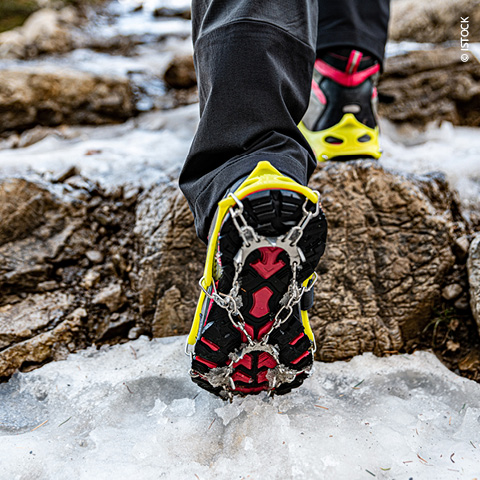 Nahaufnahme einer Person mit Eiskrallen an bunten Wanderschuhen, die über felsiges, eisiges Gelände läuft. Der Fokus liegt auf den Krallen, die auf der rutschigen Oberfläche Halt finden und zeigt die Ausrüstung für Outdoor-Winteraktivitäten.